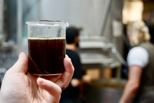 Load image into Gallery viewer, Hand holding a beaker of brown wort, with a blurred brewery in the backgrond