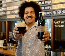 Load image into Gallery viewer, Man holding a bottle and glass of beer in a bar setting