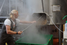 Load image into Gallery viewer, A brewer digging out brown malt from a mash tun into a green bin, smoke and steam billowing out into the brewery