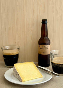 Bottle of 'The Kernel' beer with a glass of dark beer and a piece of cheese on a plate.