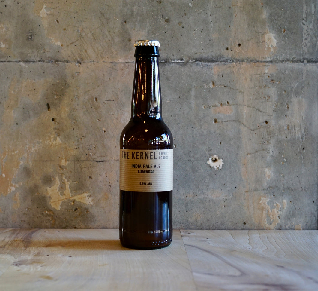 A bottle of The Kernels India Pale Ale - Luminosa on a wooden surface with a textured wall background