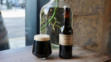 Load image into Gallery viewer, Bottle and glass of dark beer on a wooden surface with a blurred background
