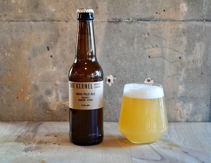 Bottle and glass of beer labeled 'The Kerne India Pale Ale - Galaxy, SImcoe, Citral' on a wooden surface with a concrete wall background.