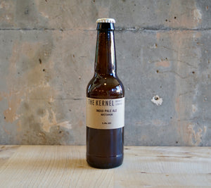 Beer bottle labeled 'The Kernel' on a wooden surface with a concrete wall background