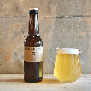 A bottle of Kernel Brewery Pils Ella stood next to a glass of golden beer infront of a concrete wall